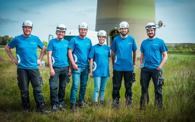 Sechs PLANETA-Mitarbeiter in Schutzkleidung stehen vor Windkraftturm – Einsatz im Feld bei Wartung und Technik